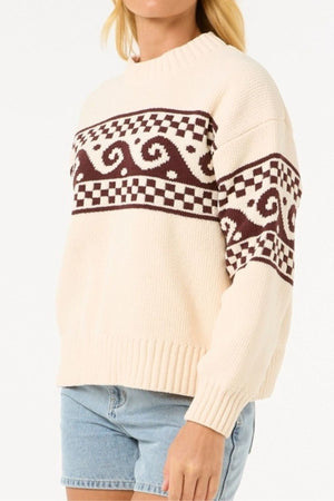 Someone wearing the Rip Curl Soleil Knit Crew—an oversized cream sweater with brown geometric and wave patterns made from recycled poly chenille—paired with light blue denim shorts.