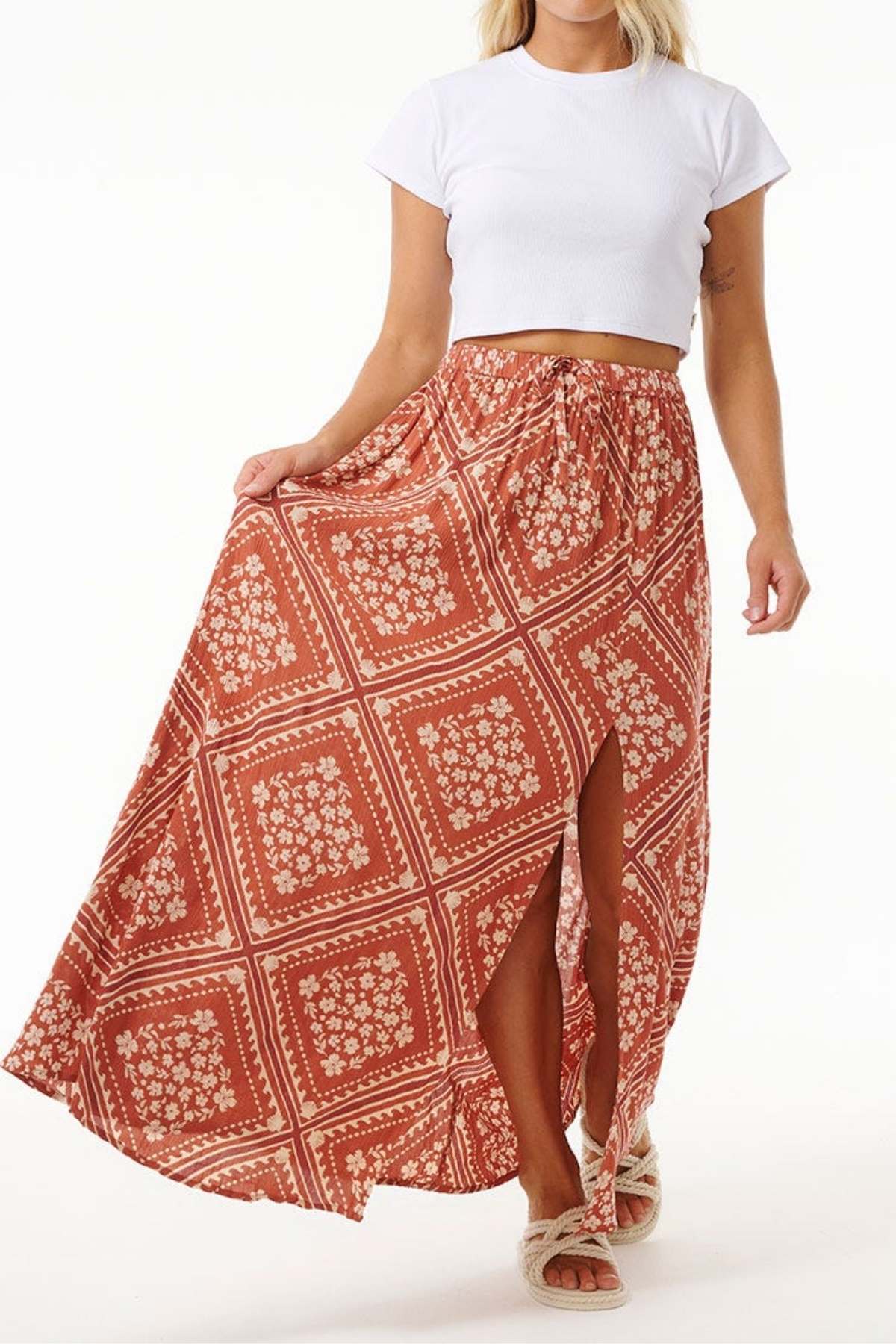 A person is wearing a white cropped t-shirt and the Rip Curl Soleil Maxi Skirt, a long, flowing red maxi skirt adorned with a floral pattern and crafted from sustainable Lenzing Ecovero. The skirt features a front slit, completing their eco-friendly look with light-colored sandals.