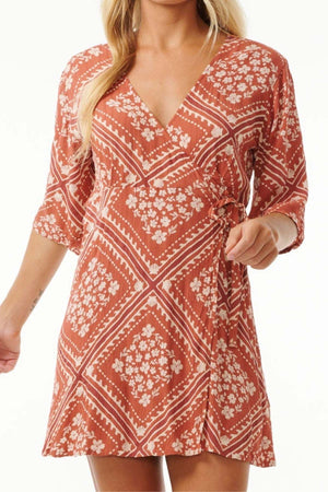 A person wearing a Rip Curl Soleil Wrap Mini Dress in coral and white floral-pattern with a V-neck and short sleeves. The dress showcases a wrap design complete with a side tie, enhancing the chic summer look. Their long, wavy blonde hair perfectly complements the ensemble.