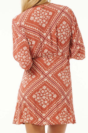 From the back, a person is seen wearing the Rip Curl Soleil Wrap Mini Dress, showcasing its vibrant red color with delicate white floral designs and diamond motifs. Crafted from eco-friendly Lenzing™ Ecovero™ fabric, this dress combines sustainability with style. The individual's blonde hair falls gracefully over the short sleeves, enhancing the dress's relaxed fit.