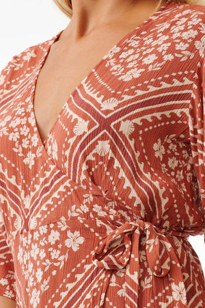 A close-up of someone wearing the rust-colored Rip Curl Soleil Wrap Mini Dress, adorned with white floral and geometric patterns. Made from Lenzing™ Ecovero™, this dress features a V-neckline, a tied waist detail, and short sleeves that emphasize its intricate designs.