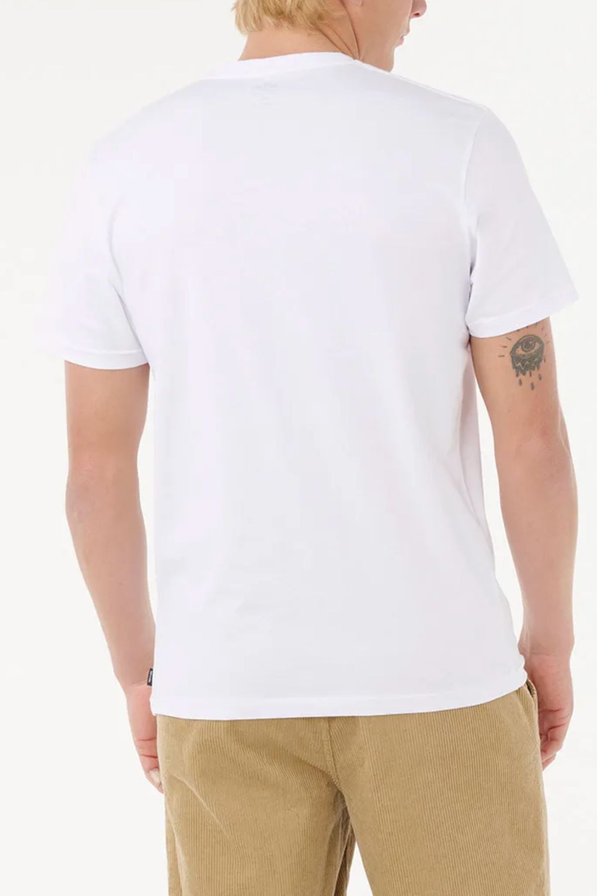 A person with light skin and blond hair is seen from the back wearing a plain white Rip Curl Staple Filler Tee and tan corduroy pants. A small eye and star tattoo is on their left arm.