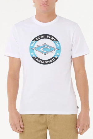 Wearing the Rip Curl Staple Filler Tee in white with a circular blue and black "Rip Curl Surf Co Established 69" chest logo and tan corduroy pants, a person stands against a plain white background.