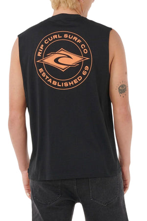 A person with short blond hair wears a black Rip Curl Staple Muscle shirt made from 100% cotton, featuring an orange "Rip Curl Surf Co Established 69" logo on the back and has a tattoo of an eye with rays on their left arm.