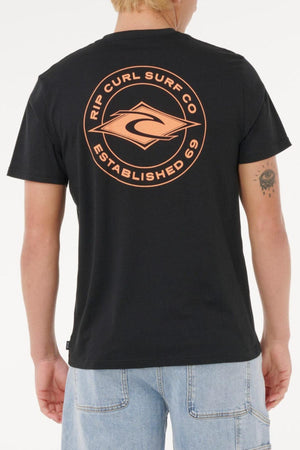 A person, back to the camera, wears a black Rip Curl Staple Tee with an orange “Rip Curl Surf Co Established 69” back logo and light blue denim shorts. The tee is made from cotton for comfort.