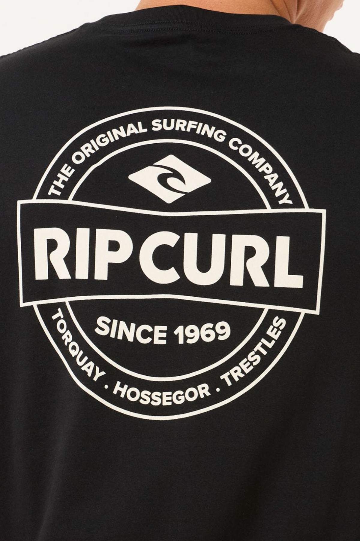 The back of the black Rip Curl Staple Tee showcases a white circular logo with the text "Rip Curl - The Original Surfing Company - Since 1969," and locations "Torquay, Hossegor, Trestles." Made from soft cotton jersey, it's ideal for casual occasions.