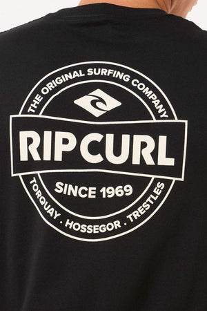 The back of the black Rip Curl Staple Tee showcases a white circular logo with the text "Rip Curl - The Original Surfing Company - Since 1969," and locations "Torquay, Hossegor, Trestles." Made from soft cotton jersey, it's ideal for casual occasions.