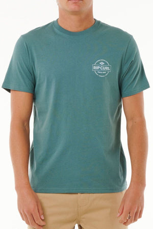 Wearing the Rip Curl Staple Tee in teal, made from soft cotton jersey with a small white logo on the left chest, and beige pants, a person stands against a plain white background.