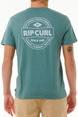 Wearing a teal Rip Curl Staple Tee in soft cotton jersey with a bold white "Rip Curl Since 1969" logo and beach locations on the back, a person faces away, revealing a tattoo on their left arm.