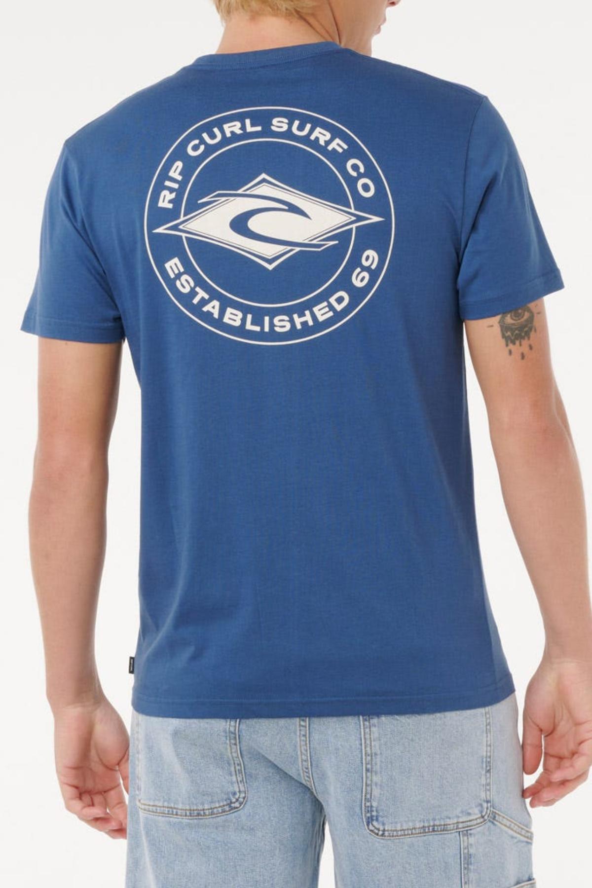 A person wearing the Rip Curl Staple Tee in blue, featuring a large white "Rip Curl Surf Co Established 69" logo on the back, is paired with light blue denim shorts. The comfortable cotton t-shirt is shown from behind.