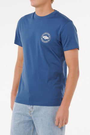 A person wears a blue Rip Curl Staple Tee featuring a small round logo on the left chest, paired with light blue jeans, and stands against a plain white background. This tee provides comfortable everyday wear.