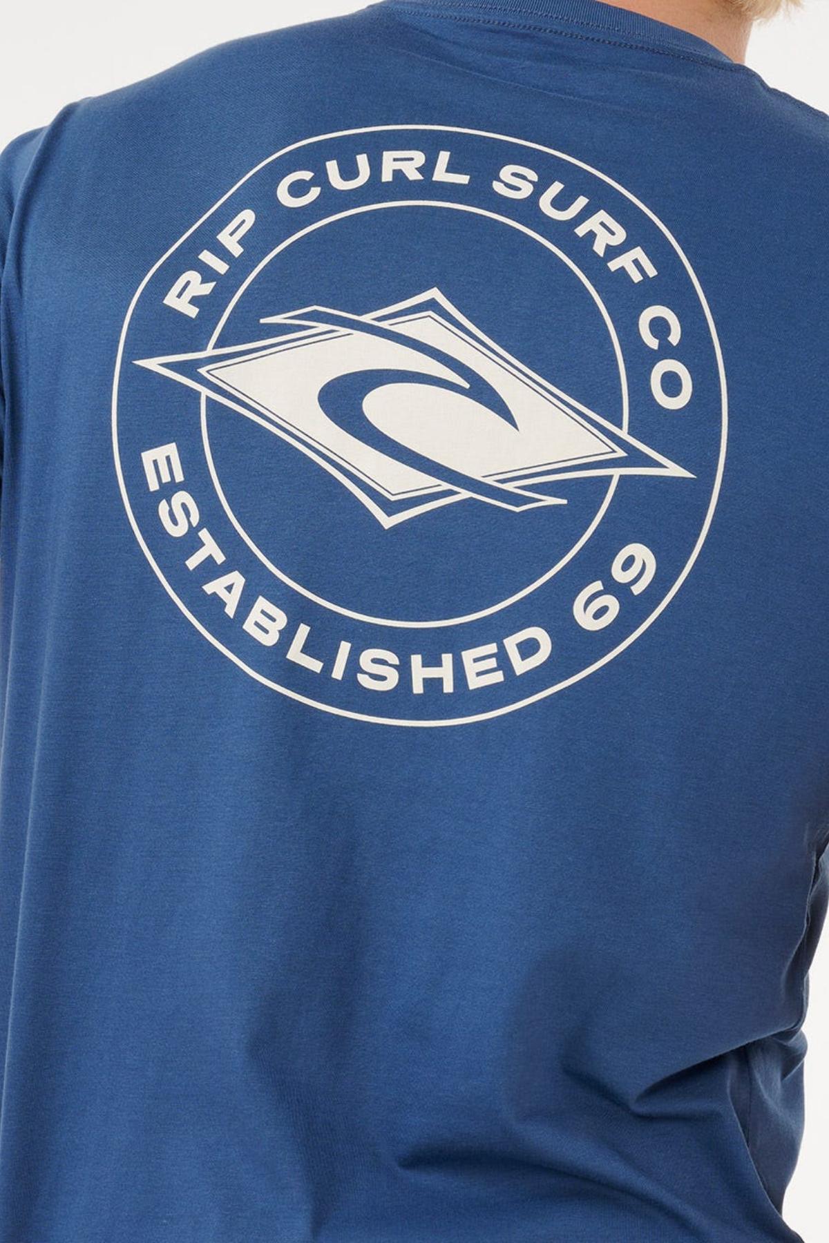 The back of the Rip Curl Staple Tee features a white circular "Rip Curl Surf Co Established 69" logo and wave graphic on blue soft cotton for a comfortable fit.