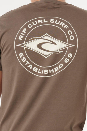 Someone wearing the Rip Curl Staple Tee in brown, featuring a large white Rip Curl Surf Co. logo, "Established 69" text, and stylized diamond-shaped waves on the back. This cotton t-shirt ensures a comfortable fit.