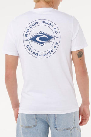 A person wears a Rip Curl Staple Tee, a white cotton jersey with a large blue back logo, styled with light blue jeans, short blonde hair, and a tattoo on their left arm.