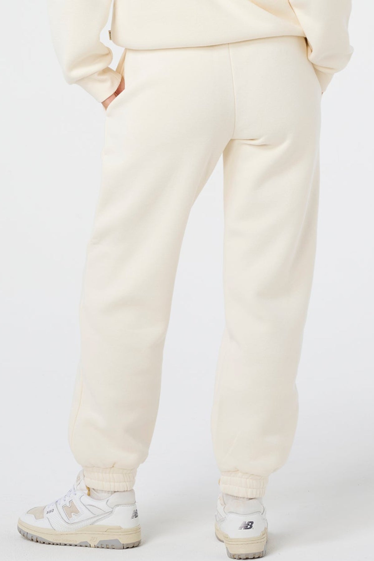A person stands with one hand in their pocket, shown from the back, wearing Rip Curl Surf Staple Trackpants and a matching cream sweatshirt with white New Balance sneakers for a relaxed surf-inspired look.