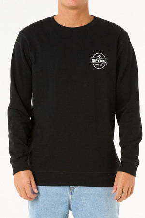 A person stands against a white background, wearing the black Rip Curl Stapler Crew sweatshirt—crafted from cotton polyester fleece with a small white logo on the upper left chest—and light blue jeans.