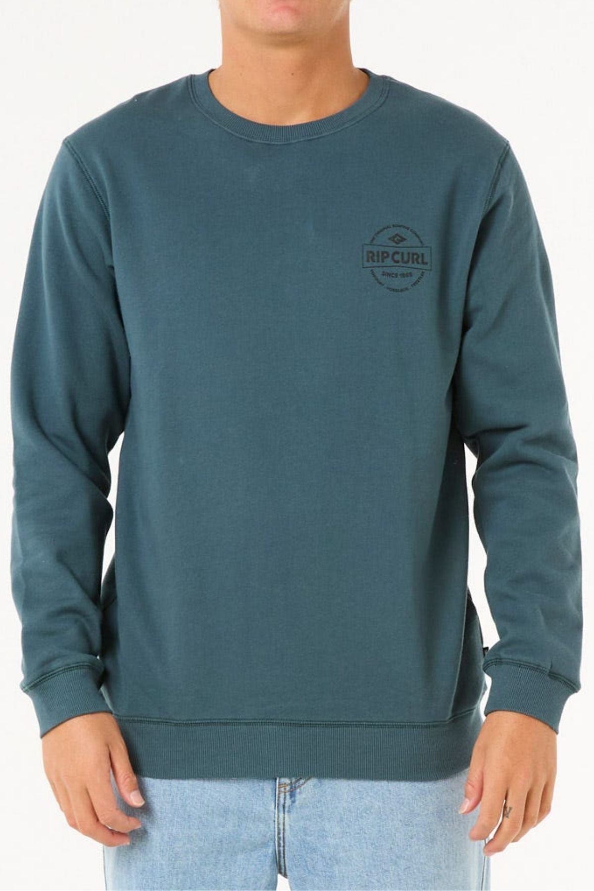 A person wears the Rip Curl Stapler Crew sweatshirt in teal, featuring soft cotton polyester fleece, paired with light blue jeans. The upper body is shown against a plain white background.