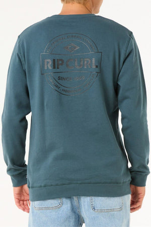 A person seen from behind wears a teal Rip Curl Stapler Crew sweatshirt with a circular logo and "RIP CURL SINCE 1969" on the back, paired with light blue jeans—ideal for adventures.