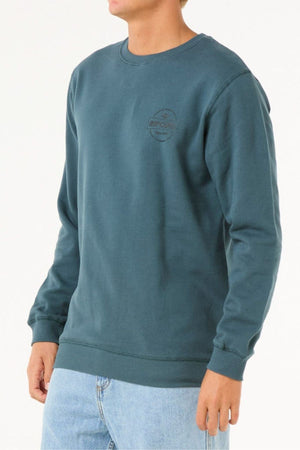 Wearing the Rip Curl Stapler Crew—a cotton-polyester fleece sweatshirt with a subtle left-chest "RIP CURL" logo—and light blue jeans, a person stands against a plain white background.