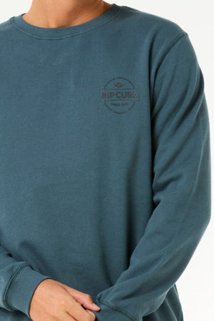 A person wears a teal Rip Curl Stapler Crew sweatshirt made from cotton-polyester fleece, with a small "Rip Curl Since 1969" logo on the left chest; their face is not visible.