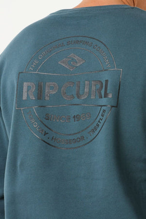 A close-up of the back of a teal Rip Curl Stapler Crew showcases the "Rip Curl" logo and text, made from soft cotton-polyester fleece for comfort and style.