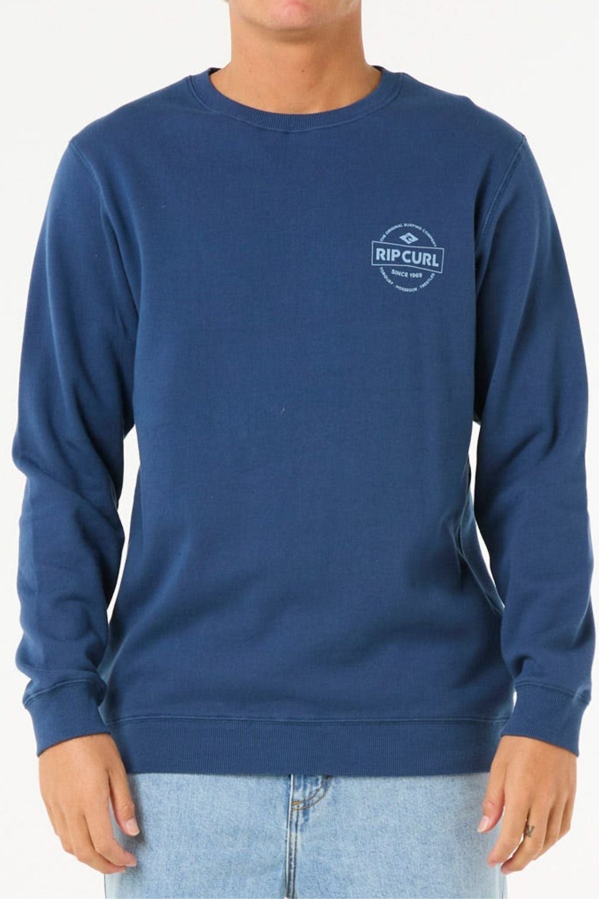 A person in the Rip Curl Stapler Crew sweatshirtโa blue cotton-poly fleece with a small white logo on the left chestโpaired with light blue jeans, stands against a plain white background.