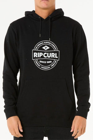 Someone wearing the Rip Curl Stapler Hood, a black cotton polyester fleece hoodie featuring a white "Rip Curl" logo plus "The Original Surfing Company," "Since 1969," and "Proudly - Hossegor - Torquay." Their face is partially obscured.