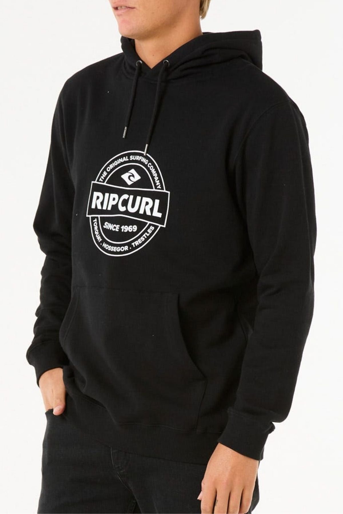 Wearing a black Rip Curl Stapler Hood hoodie with a white front logo, made of soft cotton polyester fleece, a person stands against a plain white background with one hand in the pocket.