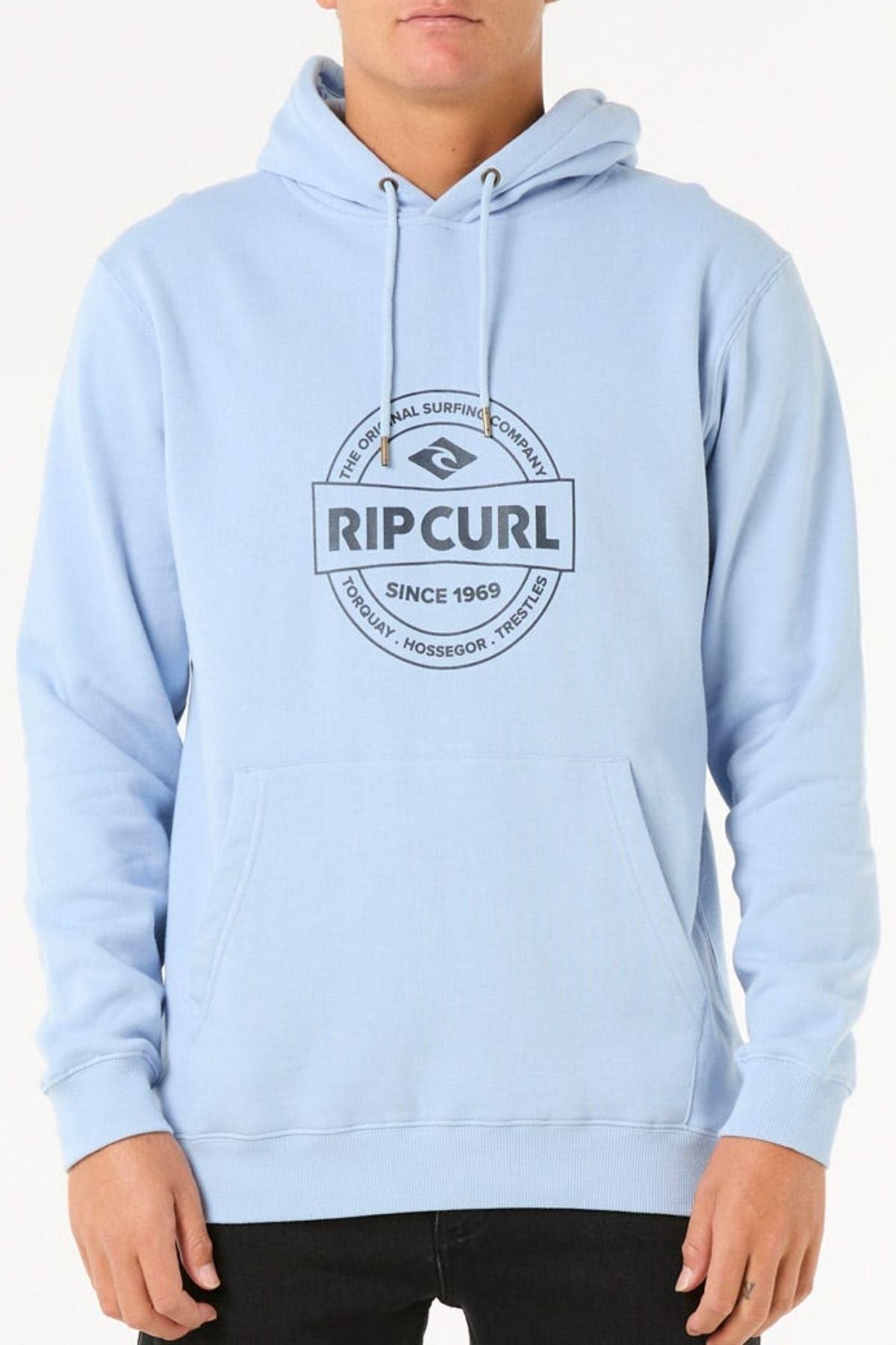 A person wears the Rip Curl Stapler Hood hoodie in light blue, made of cotton-polyester fleece with a front pocket, drawstrings, and a black circular "Rip Curl Since 1969" logo; their hands are by their sides.