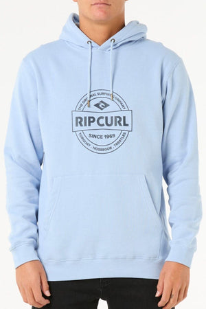 A person wears the Rip Curl Stapler Hood hoodie in light blue, made of cotton-polyester fleece with a front pocket, drawstrings, and a black circular "Rip Curl Since 1969" logo; their hands are by their sides.