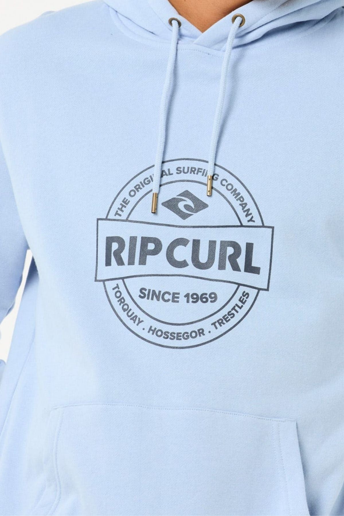 Someone wearing the Rip Curl Stapler Hood—a light blue cotton-polyester fleece hoodie—features a circular logo with "RIP CURL The Original Surfing Company Since 1969" and place names Torquay, Hossegor, and Trestles on the front.