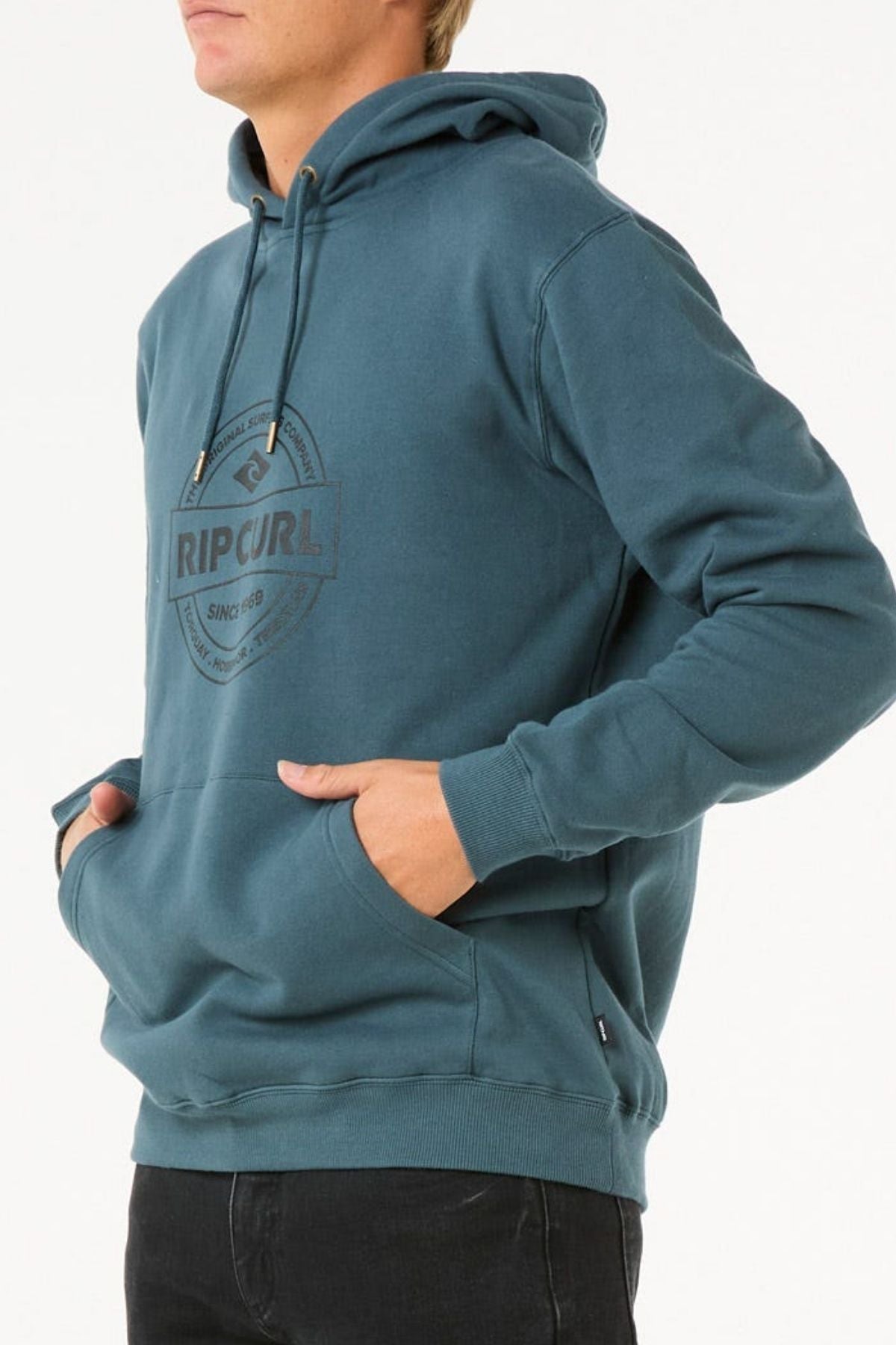 Someone wearing the Rip Curl Stapler Hood in teal, crafted from soft cotton and polyester fleece with a front pocket, pairs it with black pants and stands hands-in-pocket against a plain white background.