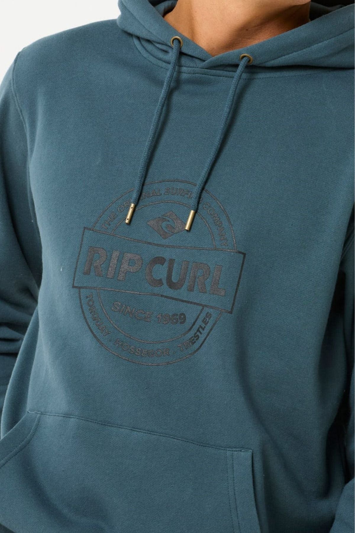 Someone wearing the Rip Curl Stapler Hood—a relaxed-fit teal hoodie crafted from soft cotton-polyester fleece, with drawstrings, a kangaroo pocket, and a circular "RIP CURL" logo on the chest.