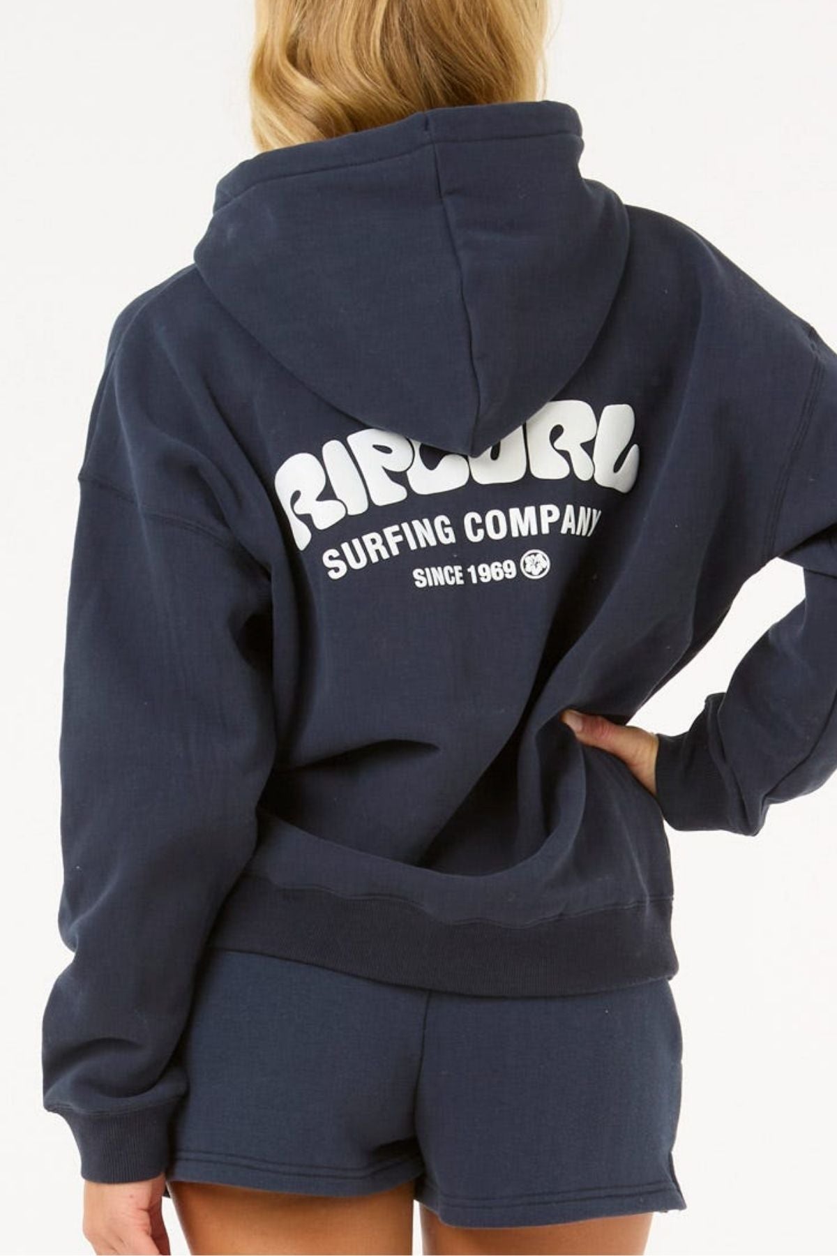 A person with long blonde hair wears a navy Rip Curl Surf Puff Heritage Hood, featuring "RIP CURL SURFING COMPANY SINCE 1969" in white on the back, with matching shorts and one hand on their hip.