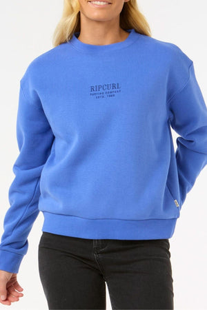 Wearing a bright blue Rip Curl Surf Staple Relaxed Crew made from cotton-polyester fleece, paired with black pants, a person stands against a plain white background. "Rip Curl Surfing Company" is printed in small text on the chest.