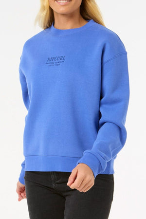 A person in a bright blue Rip Curl Surf Staple Relaxed Crew sweatshirt and black pants smiles while standing against a plain white background.