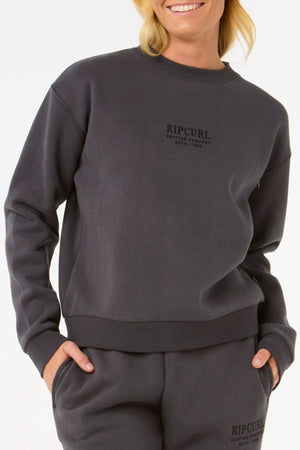 A person smiles with one hand in a pocket, wearing the Rip Curl Surf Staple Relaxed Crew and matching sweatpants in dark gray fleece, standing out against a plain white background.