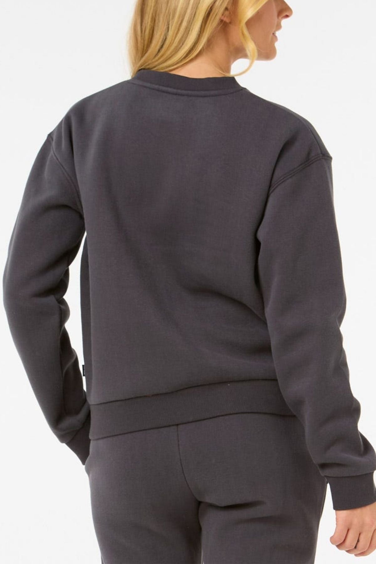 A person with long blonde hair, seen from the back, wears the Rip Curl Surf Staple Relaxed Crew in dark gray cotton-polyester fleece with matching pants against a plain white background.
