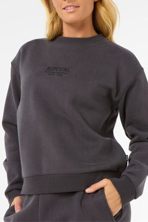 A woman with blonde hair poses casually in a dark gray Rip Curl Surf Staple Relaxed Crew, a cotton polyester fleece sweatshirt, standing with one hand in her pocket against a plain white background.