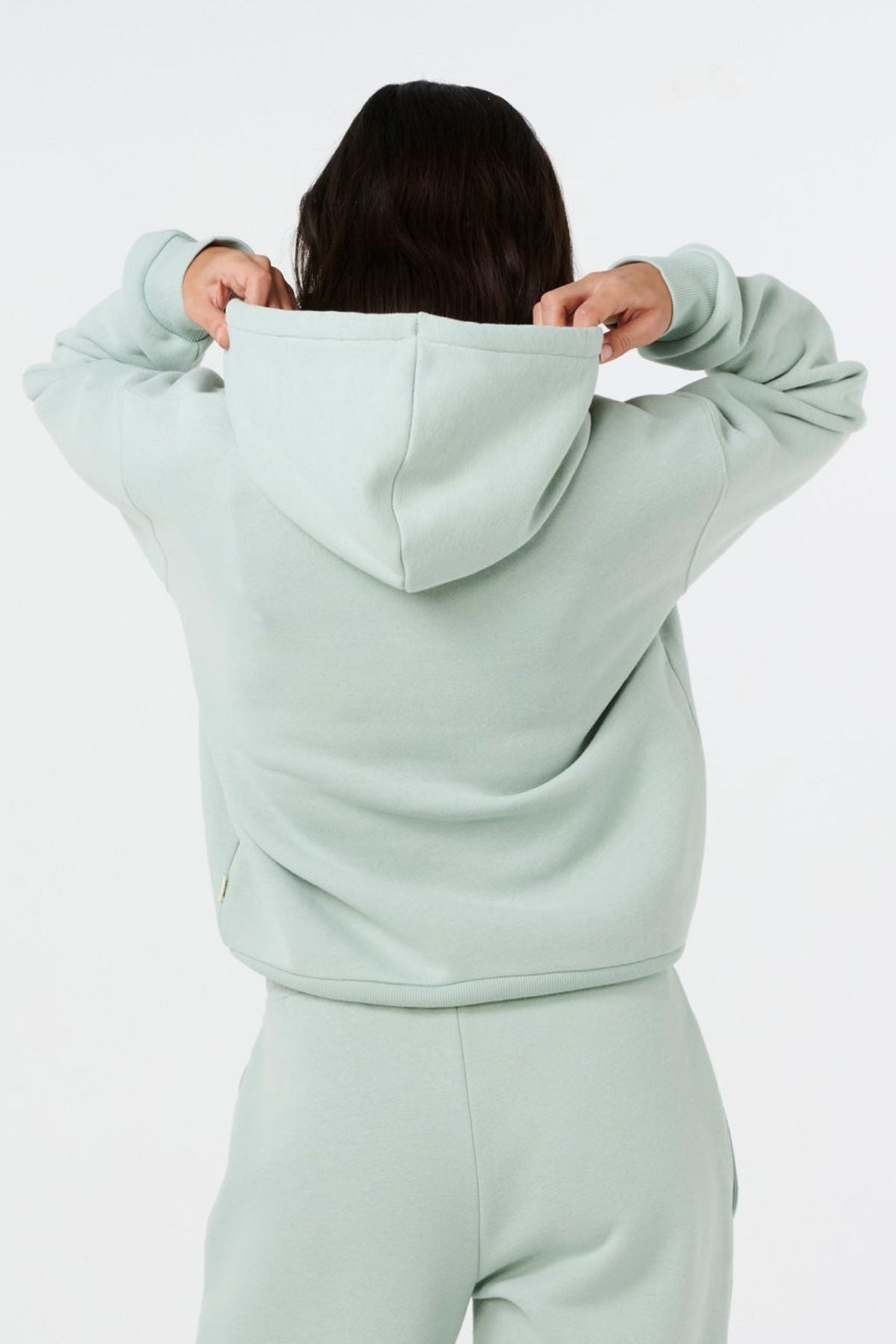 A person is seen from the back, hands lifting the hood of a light green Rip Curl Surf Staple Relaxed Hood. The relaxed-fit pullover stands out against a plain white background.