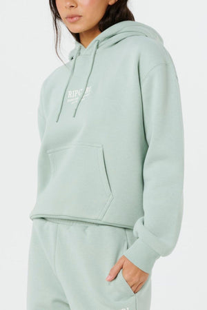 Wearing the Rip Curl Surf Staple Relaxed Hood in light green with a front pocket and matching sweatpants, this person poses casually against a white background—a laid-back essential for surf-inspired comfort.