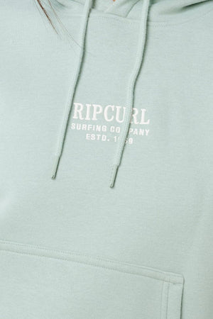 A close-up of the Rip Curl Surf Staple Relaxed Hood in light green, featuring white "RIP CURL SURFING COMPANY ESTD. 1969" text on the chest, a front pocket, drawstrings, and a relaxed fit—an essential surf pullover.