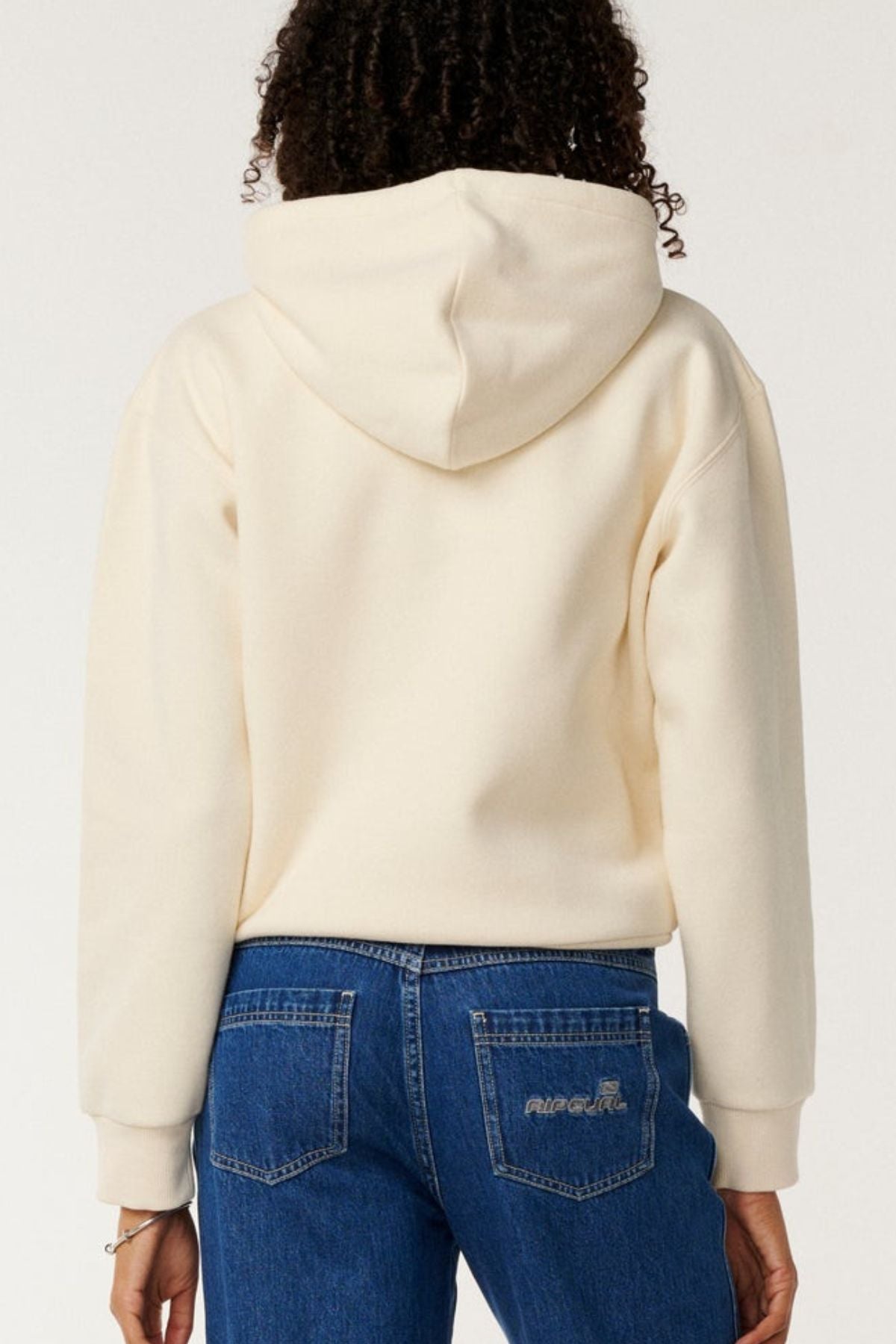 A person with curly hair is shown from the back, wearing a cream Rip Curl Surf Staple Relaxed Hood and blue jeans. The relaxed fit pullover has the hood up, and there’s embroidered text on one back pocket of the jeans.