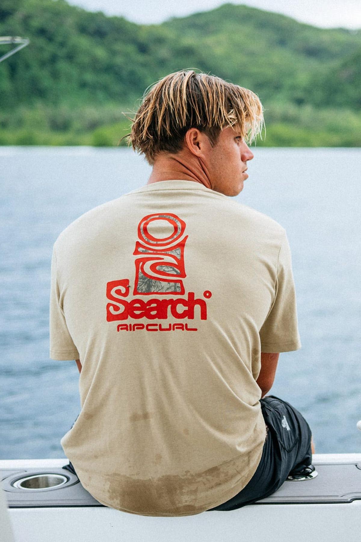 A young man with wet blond hair sits on a boat’s edge, facing away, wearing a beige Rip Curl Vaporcool Search Stack Tee with a red logo. Lush green hills and calm water can be seen in the background.