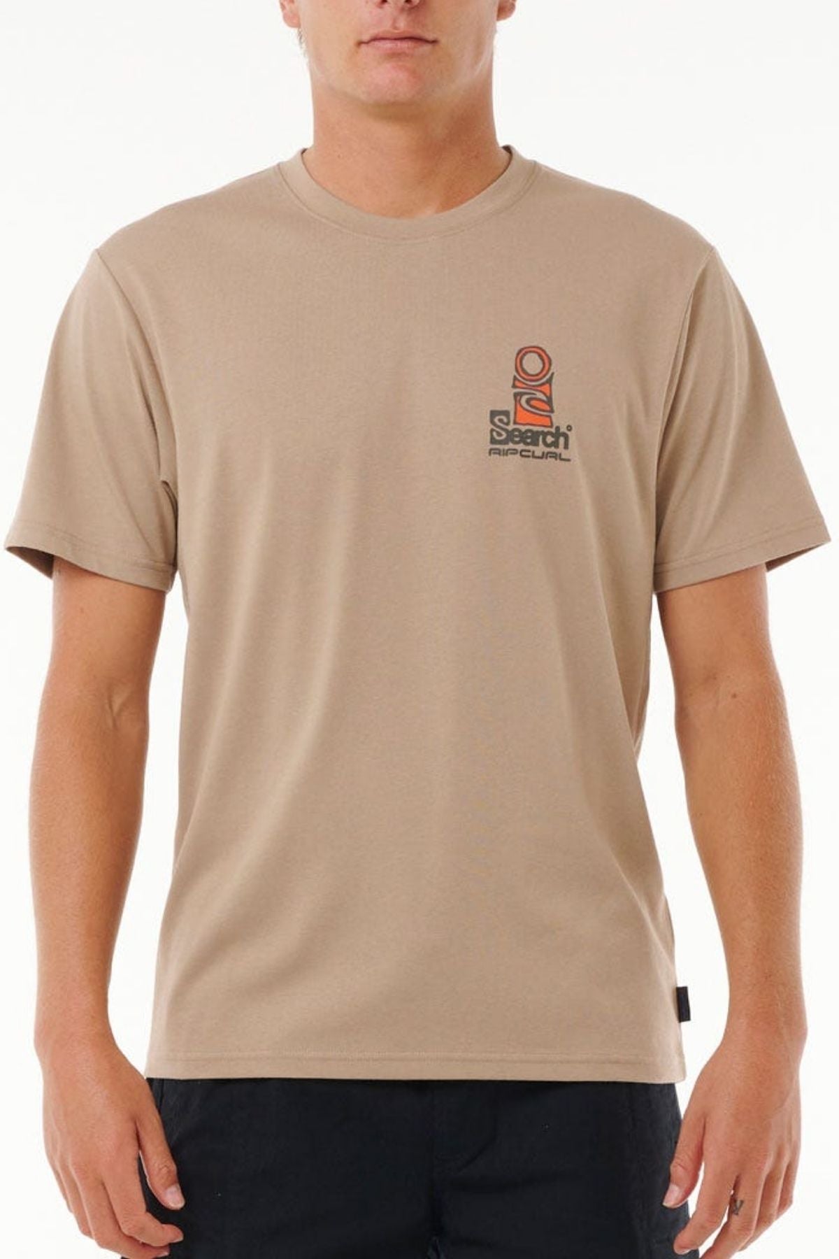 A person wearing the Rip Curl Vaporcool Search Stack Teeβa beige short-sleeve t-shirt with a small left chest logo and quick-dry Vaporcool techβpaired with black pants, stands against a plain white background.
