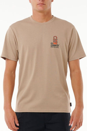 A person wearing the Rip Curl Vaporcool Search Stack Teeβa beige short-sleeve t-shirt with a small left chest logo and quick-dry Vaporcool techβpaired with black pants, stands against a plain white background.