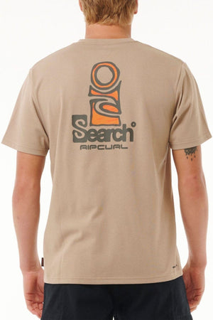 Someone stands against a plain white background wearing the Rip Curl Vaporcool Search Stack Tee, a beige shirt featuring a large orange and black graphic and βSearch Ripcurlβ printed on the back, made with quick-dry Vaporcool technology.