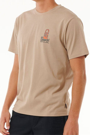 Model wears the Rip Curl Vaporcool Search Stack Tee in beige, a short-sleeve shirt with a small chest logo and quick-dry Vaporcool technology for comfort. Paired with dark pants, set against a plain, light background.