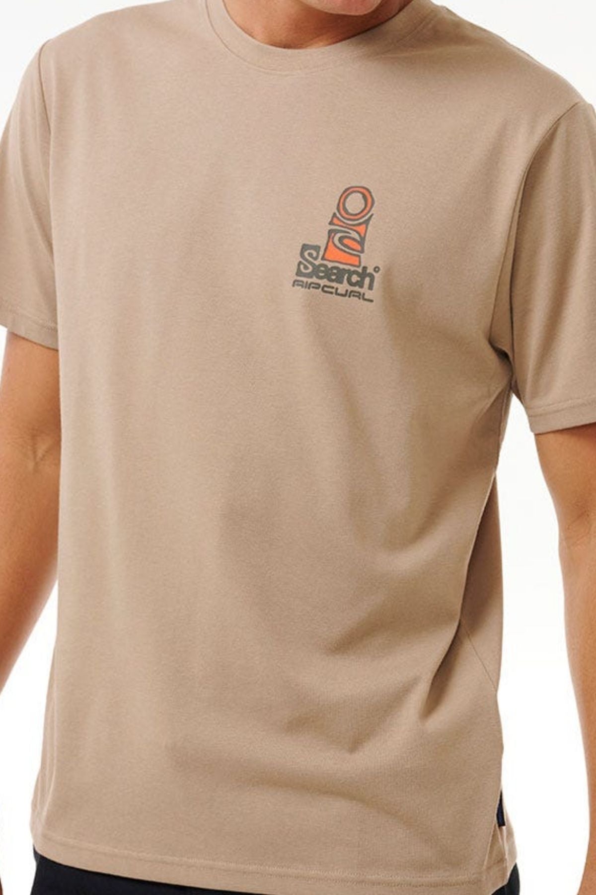 A person wears the Rip Curl Vaporcool Search Stack Tee, a beige relaxed-fit short-sleeved shirt with a small multicolored "Search Rip Curl" logo on the left chest. The person's head is not visible in the image.