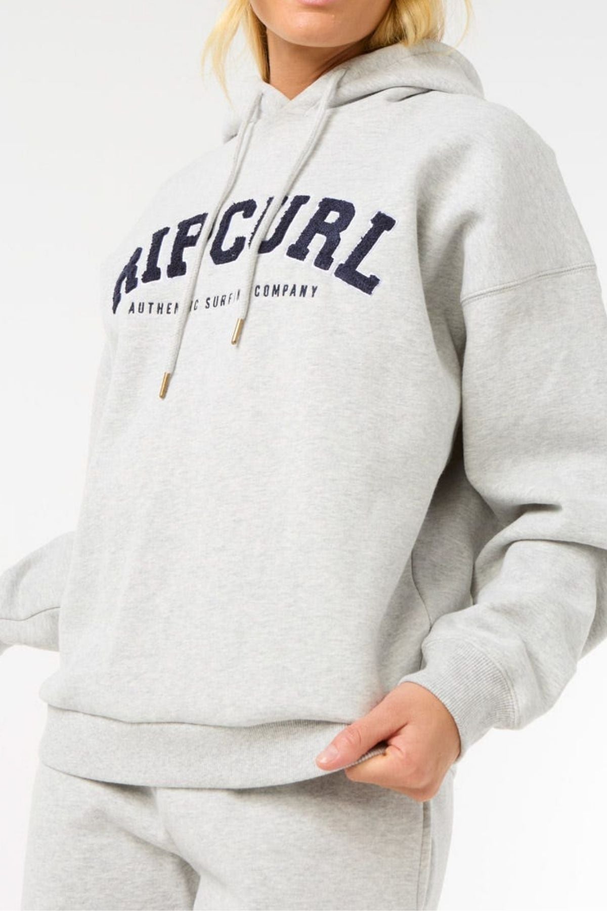 A person, face partially out of frame, wears a light gray Rip Curl Varsity Hoodie made from soft Cotton Poly Fleece with an adjustable drawcord and "RIP CURL AUTHENTIC SURF CO." in navy across the chest.